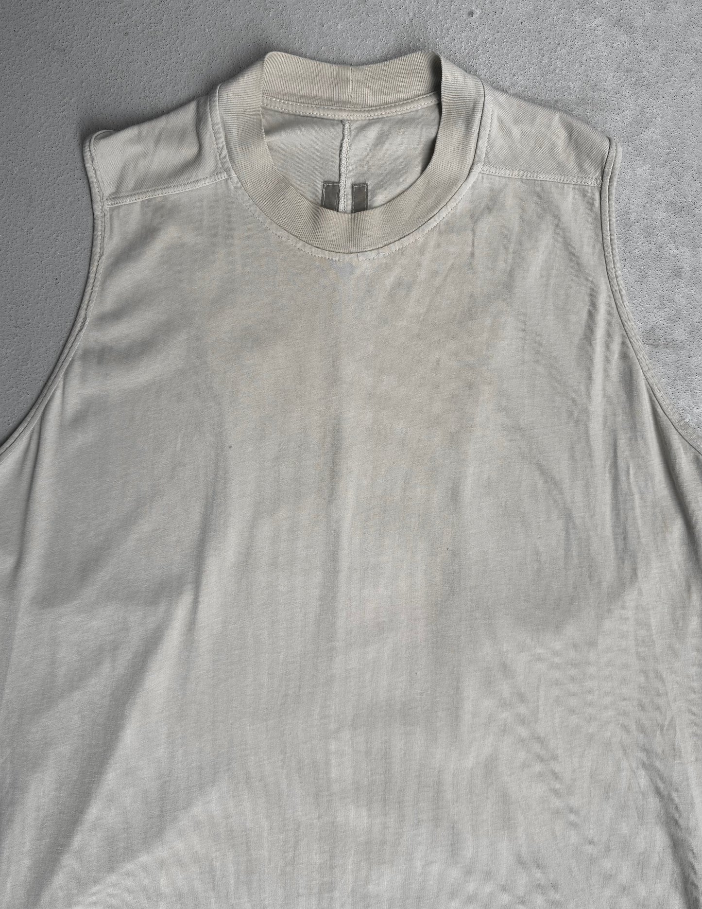 The Rick Owens DRKSHDW AW18 “SISYPHUS” Pearl Racer Tank Top, a sleeveless light beige crew neck with visible stitched seams, is displayed flat on a textured grey surface, echoing the brand’s gothic grunge aesthetic.