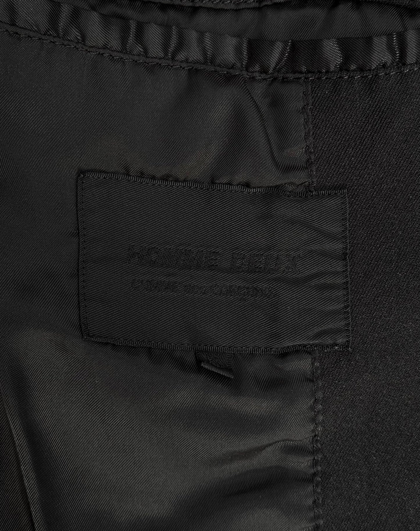 Close-up of a black clothing tag on dark fabric, subtly embroidered with text, hinting at its avant-garde fashion roots, reminiscent of iconic menswear labels like the COMME des GARÇONS HOMME DEUX Early 00s Reversible Button-up Coat by COMME des GARÇONS.
