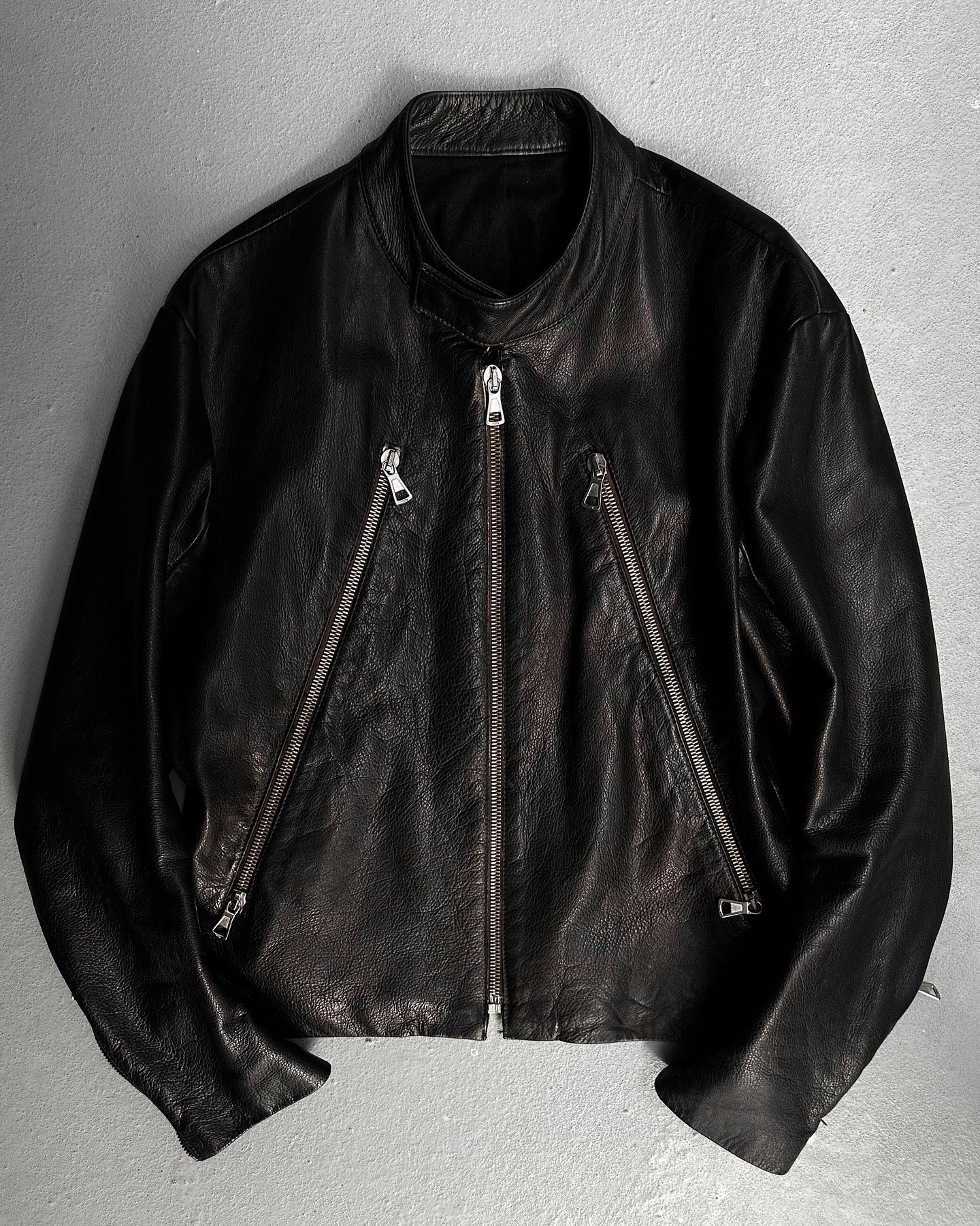 The Maison Martin Margiela AW00 Black Cowhide Leather 5-zip Rider Jacket features a stand collar, asymmetrical zip front, and zippered side pockets, shown flat on a light gray textured surface.