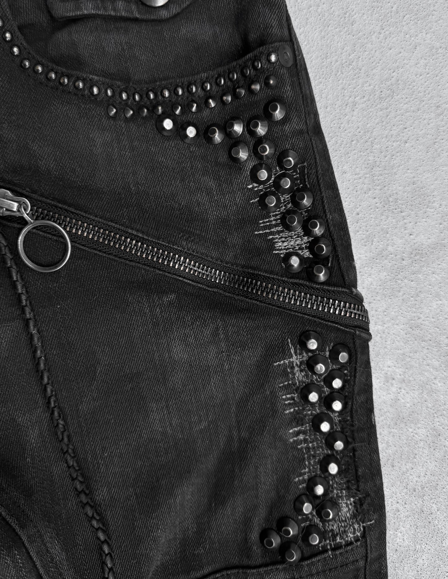 Close-up of KMRii Early 00s Goatskin Patches Studded Crush Flare-zip Denim: black denim with fading, diagonal zipper, decorative studs, distressed details. Textured surface in black and white. Fits US 29-30 waist or size 2.
