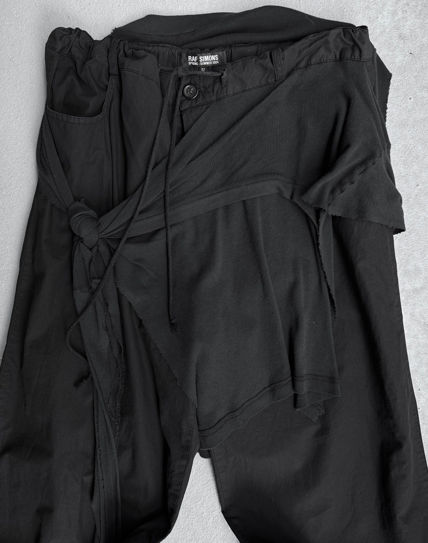 A pair of RAF SIMONS SS04 “May the Circle Be Unbroken” Reconstructed ‘Zombie’ Wide Leg Pants with asymmetrical wrap detail and waist tie, reflecting poetic minimalism, displayed on a light gray surface.