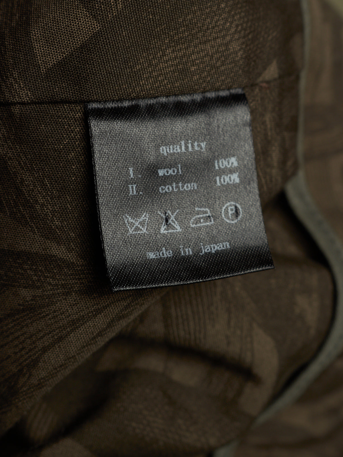 Close-up of a fabric care label sewn onto a brown textured garment. The label reads 'quality' with wool 100%, cotton 100%, and care symbols, plus 'made in japan'.