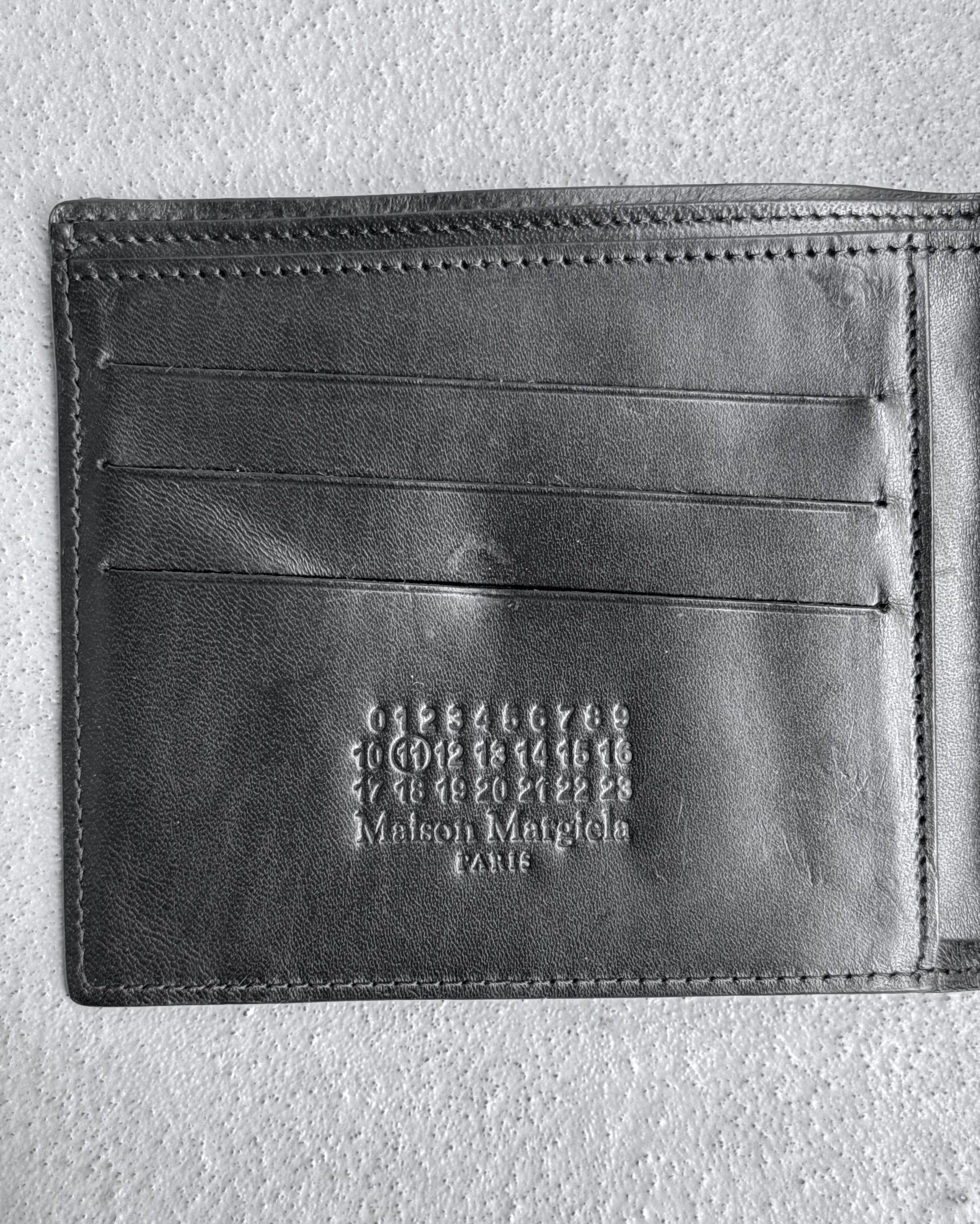 A Maison Martin Margiela AW15 Distressed Calfskin Leather Bi-fold Wallet, black, OneSize, in slightly used (8/10) condition is open to show card slots, embossed numbers, and branding on a textured gray surface.