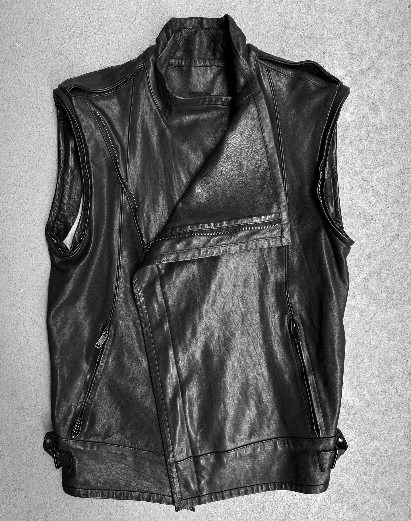 The Gareth Pugh AW09 Detachable Sleeves Horse Leather Riders Jacket, a sleeveless black piece with asymmetric front zip, wide lapel, side pockets, and adjustable hem straps, is displayed flat on a light gray surface.