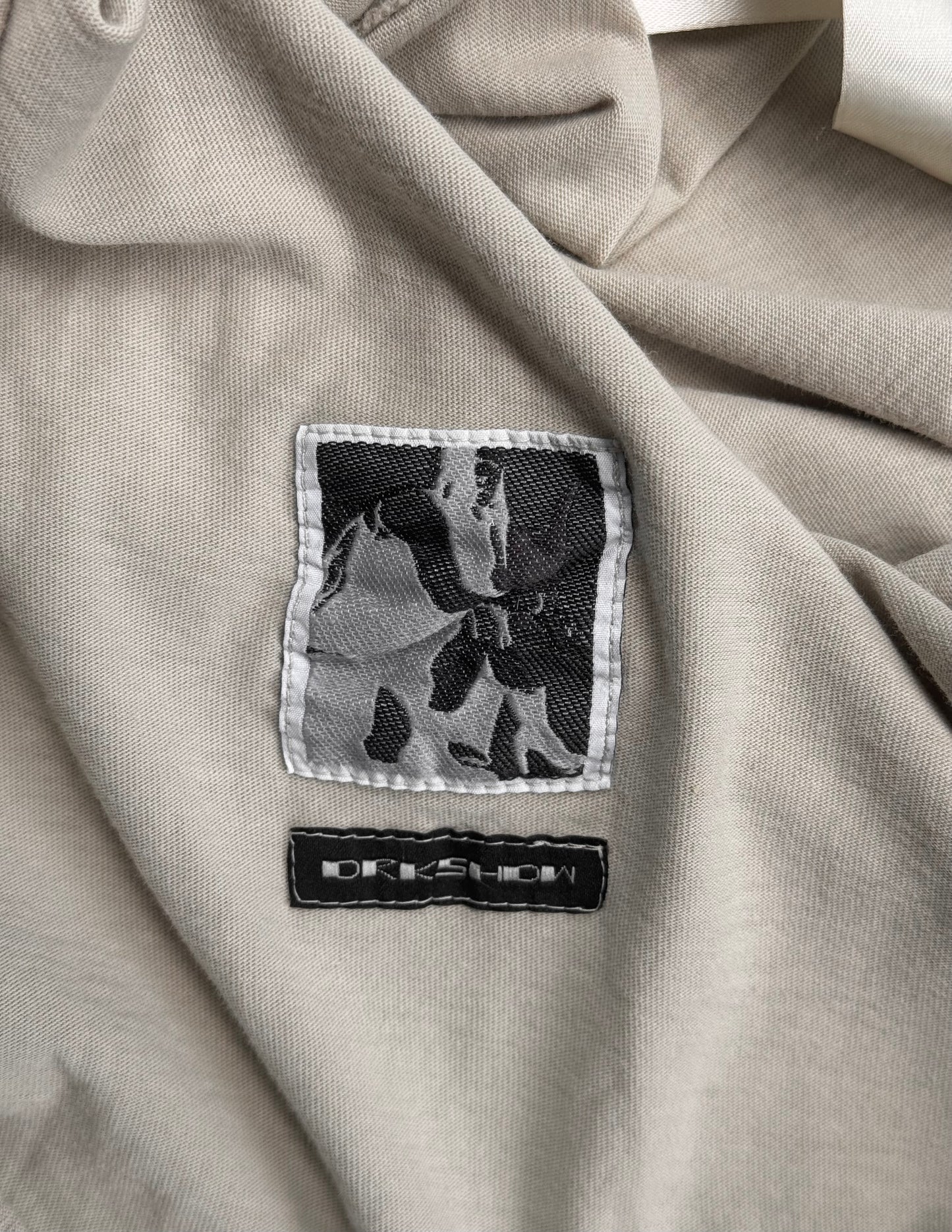 A close-up of the beige fabric from the Rick Owens DRKSHDW AW18 “SISYPHUS” Pearl Racer Tank Top shows two stitched labels: one with a black and white abstract design, the other featuring DREWSHOW in white on black.