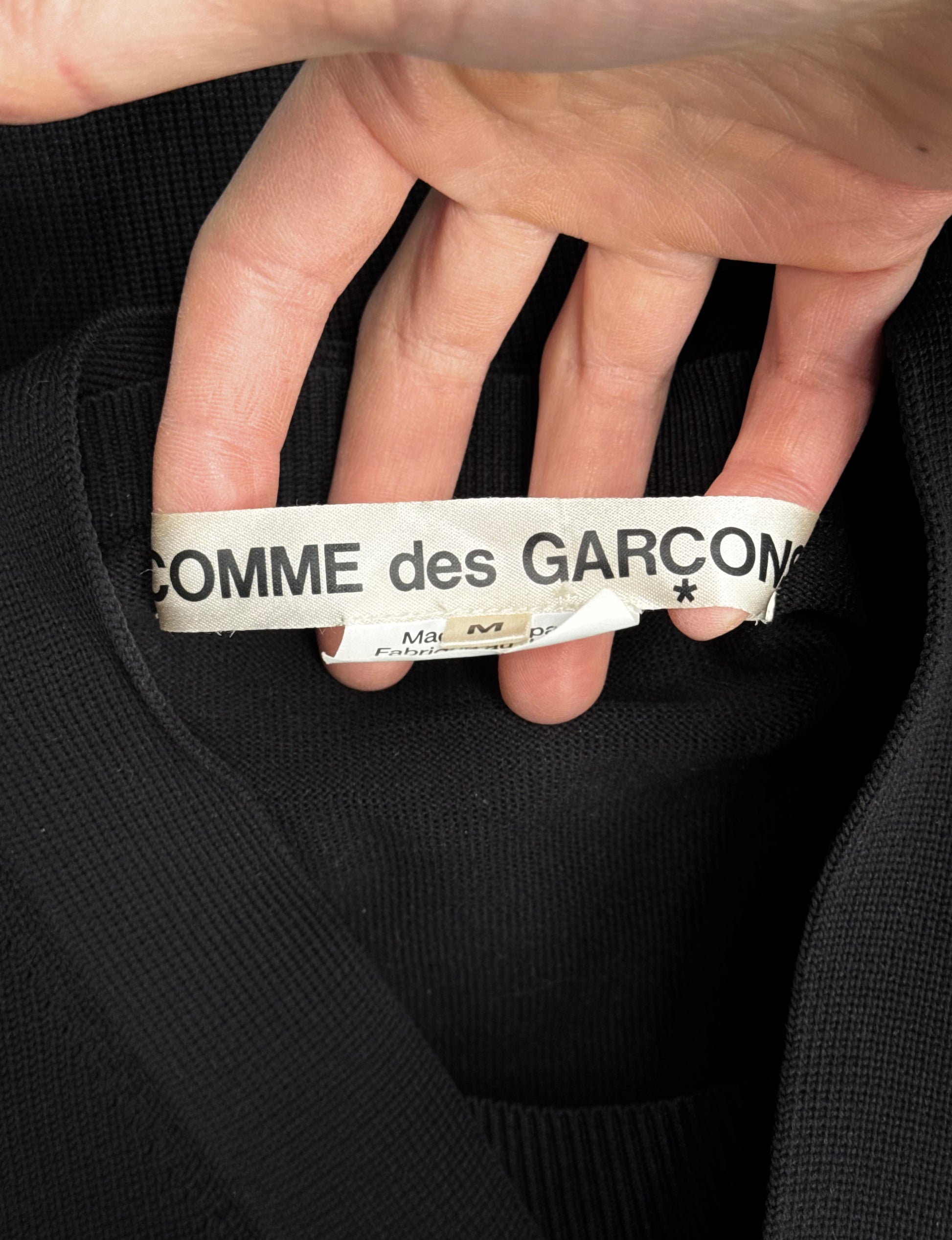 A hand reveals a slightly crumpled clothing label inside a black garment. The label reads COMME des GARÇONS SS19 Deconstructed 2-in-1 Cardigan, suggesting an avant-garde piece fit for Dover Street Markets curated collection.