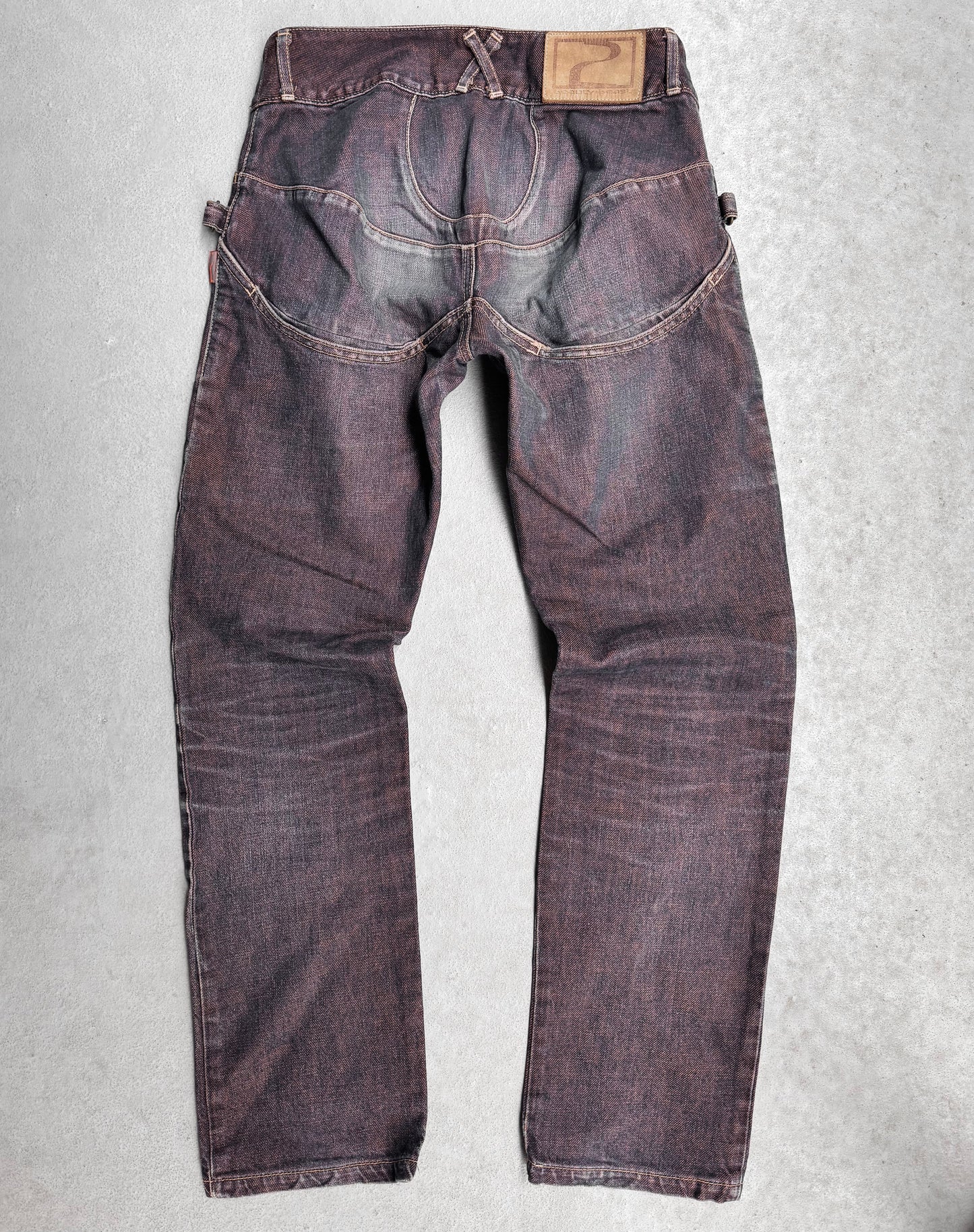Laid flat on a light gray surface, the Beauty:Beast Late 90s Over-dyed Articulate Bootcut Denim jeans feature a worn, dark blue look with fading and whiskering that capture Tokyo fashions essence. A brown leather patch adorns the waistband, and distinctive stitching enhances the rear pockets for an avant-garde flair.