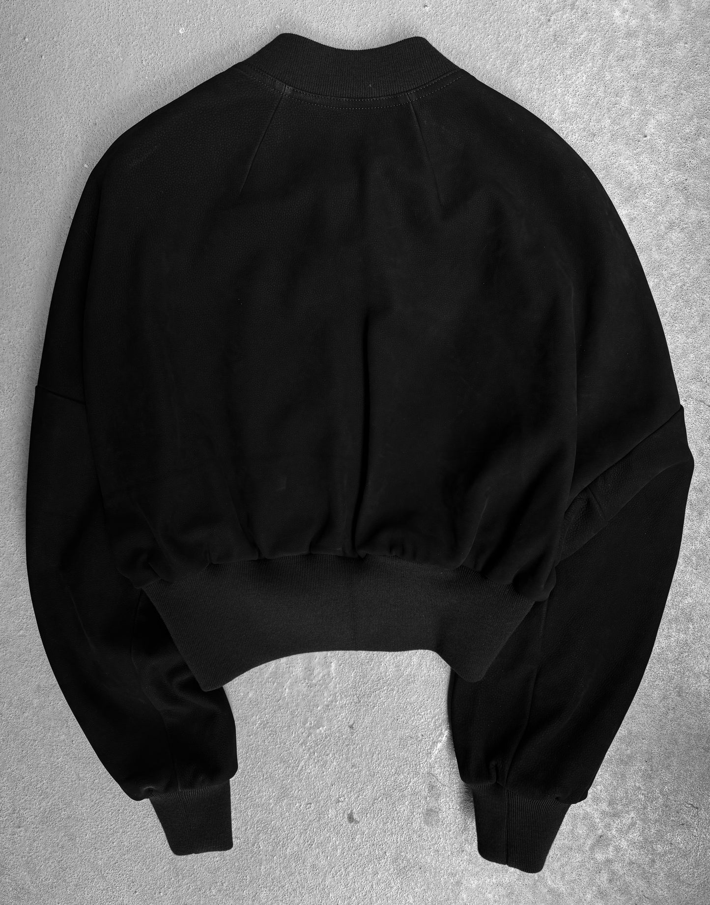 The Rick Owens SS24 “LIDO” Nubuck Leather Cropped Flight Bomber Jacket is shown laid flat, back view, on a light gray textured surface, highlighting its ribbed cuffs and waistband in true avant-garde style.