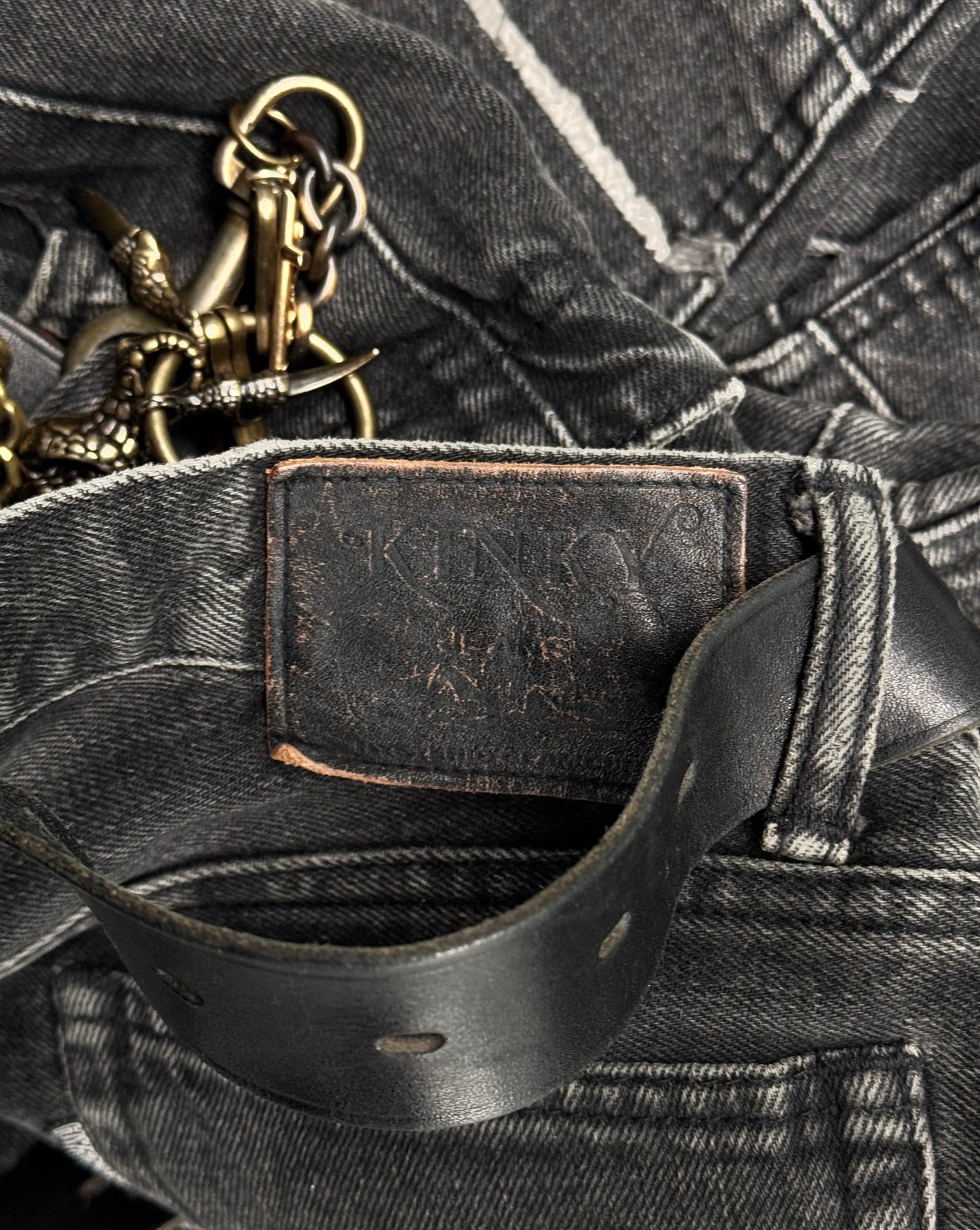 Close-up of Hysteric Glamour Early 00s Kinky Grey Flare Hagi Denim with a visible leather label partially obscured. A black worn leather belt loops through the waist, exuding punk rock vibes. A metal keychain with intricate designs is also attached to the jeans.