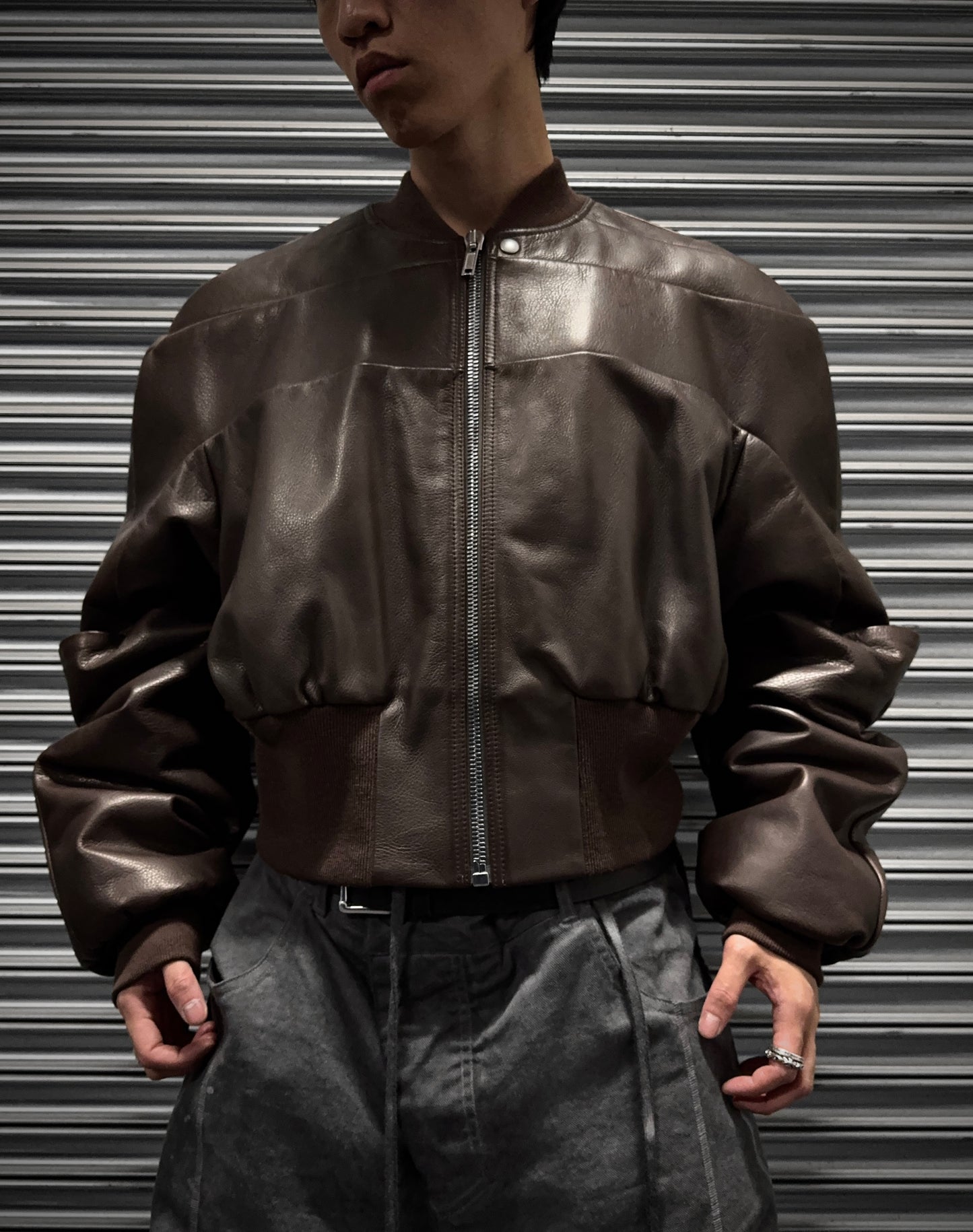 A person in Rick Owens AW23 LUXOR Girdered Leather Bomber Jacket and gray pants stands before a metallic ribbed backdrop, hands at their sides, capturing gothic grunge vibes with their face partially out of frame.