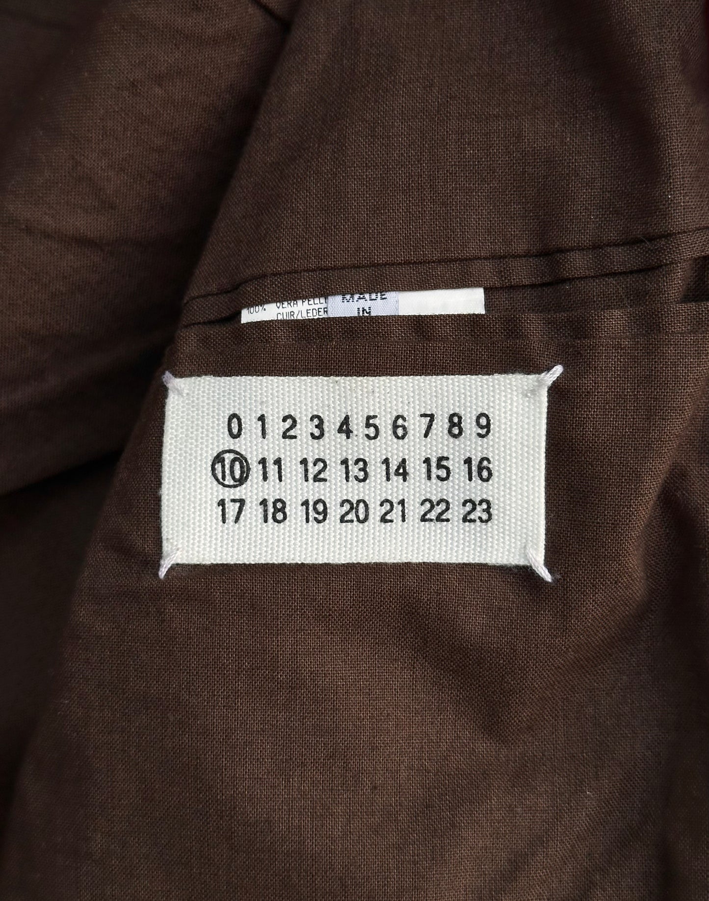 A white fabric label with black numbers 0–23 is sewn onto red cowhide leather on the Maison Martin Margiela AW00 5-zip Rider Jacket; the number 10 is circled, and a small white tag with partial text sits above.