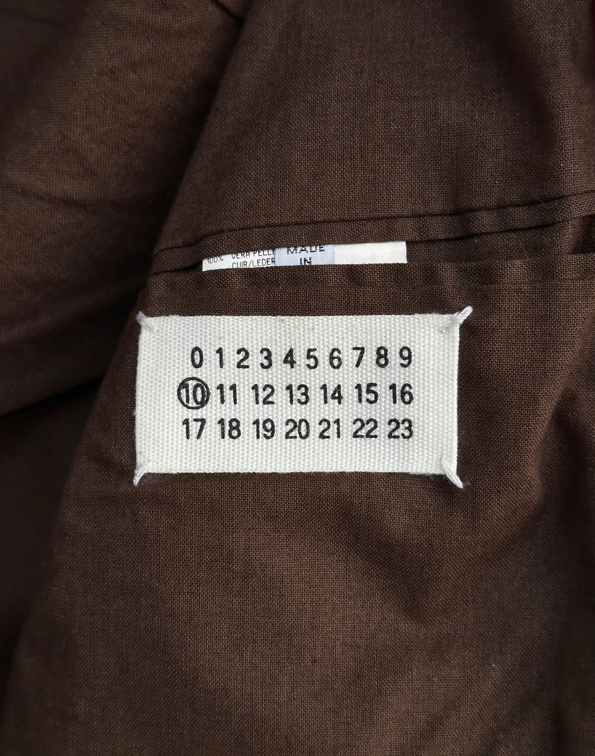 A white fabric label with black numbers 0–23 is sewn onto red cowhide leather on the Maison Martin Margiela AW00 5-zip Rider Jacket; the number 10 is circled, and a small white tag with partial text sits above.
