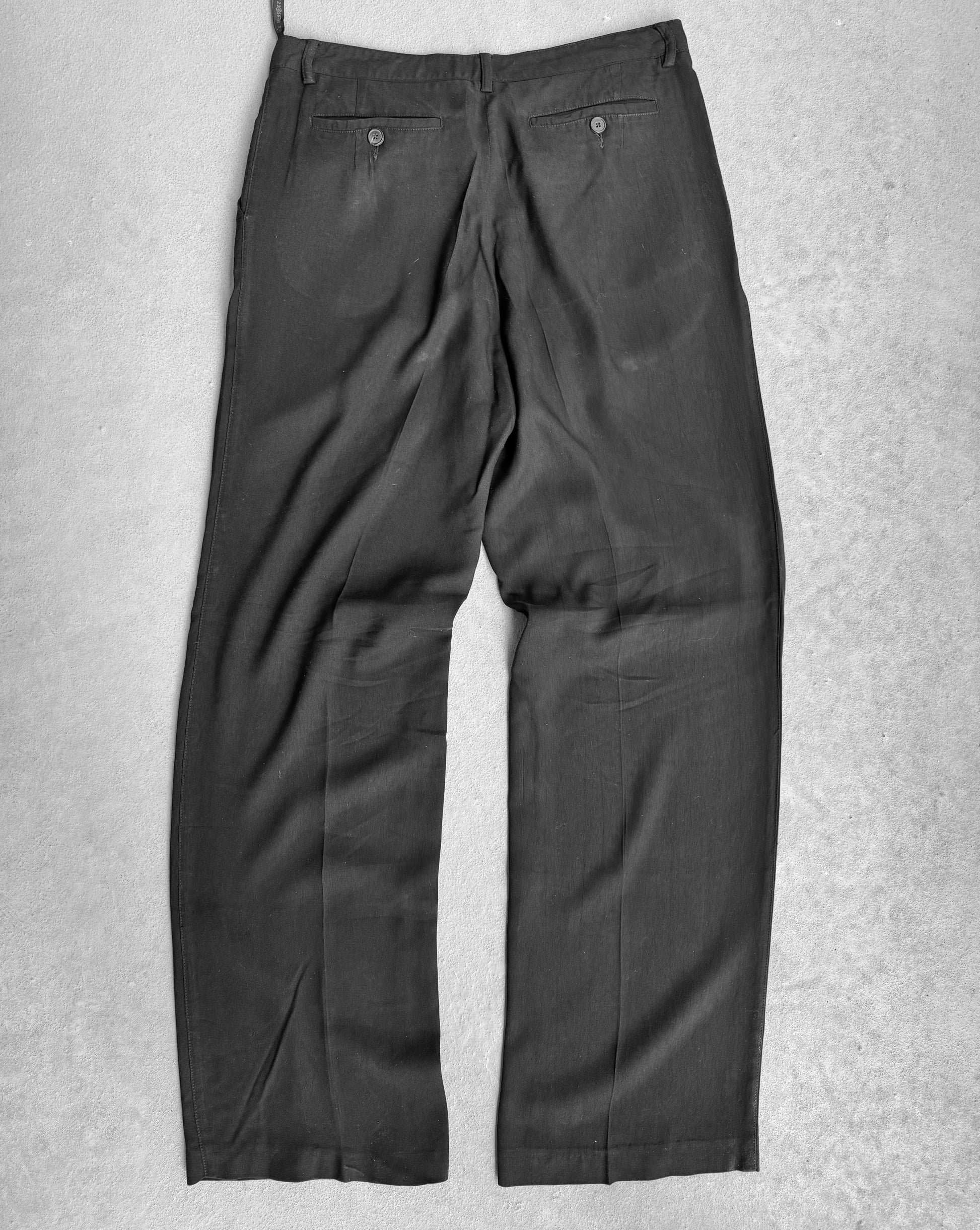 The Maison Martin Margiela Early 00s Double Pleated Dress Pants, seen laid flat on a gray surface from the back, embody avant-garde fashion with two buttoned pockets and subtle leg creases that highlight a minimalist design defying conventional branding norms.