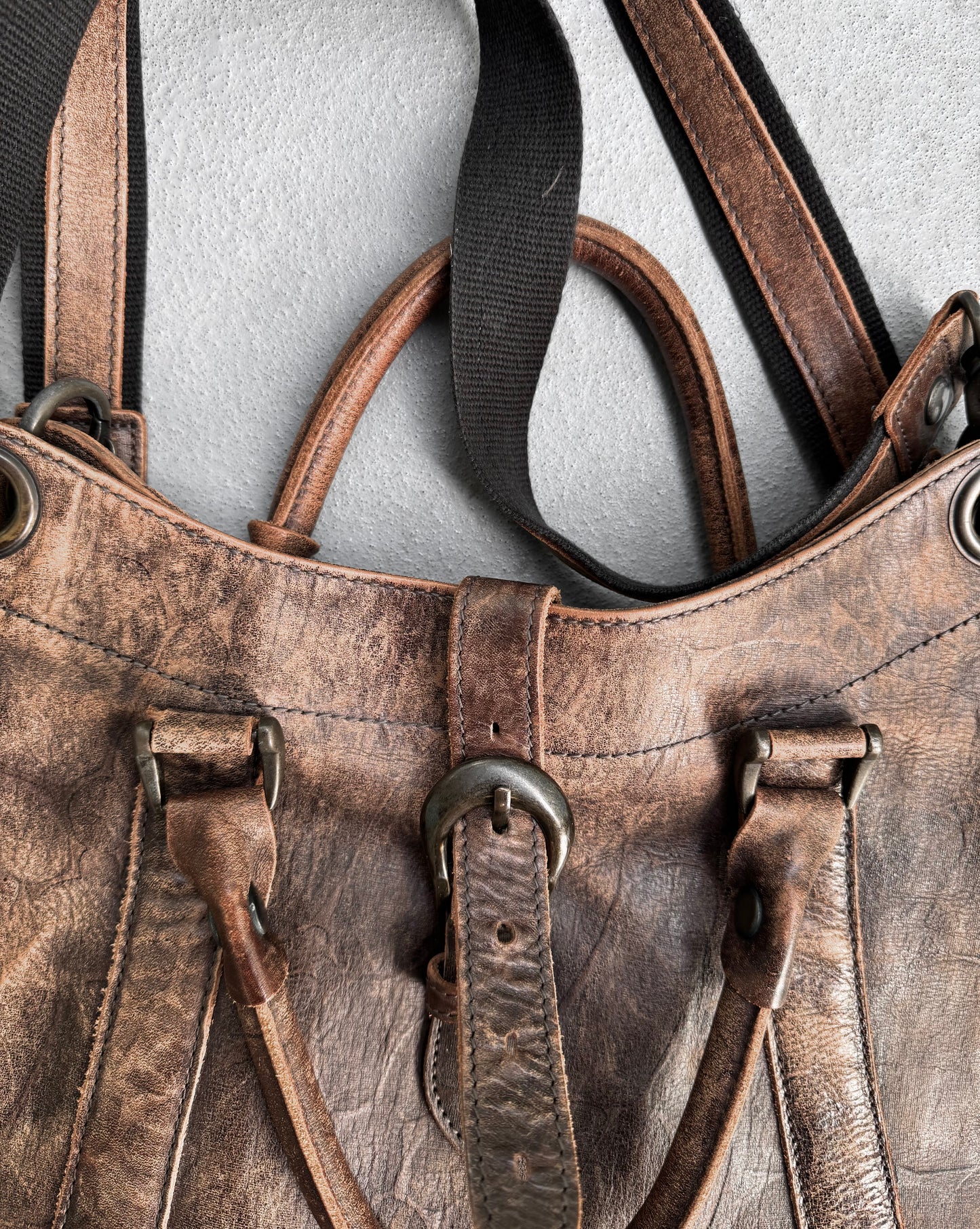 A close-up of the alfredoBANNISTER Early 00s Faded Brown Cowhide Tote Bag, with black straps on a gray background. Crafted from Japanese leather, its brass rings, central buckle, and visible stitching blend rugged charm with a vintage yet modern vibe.