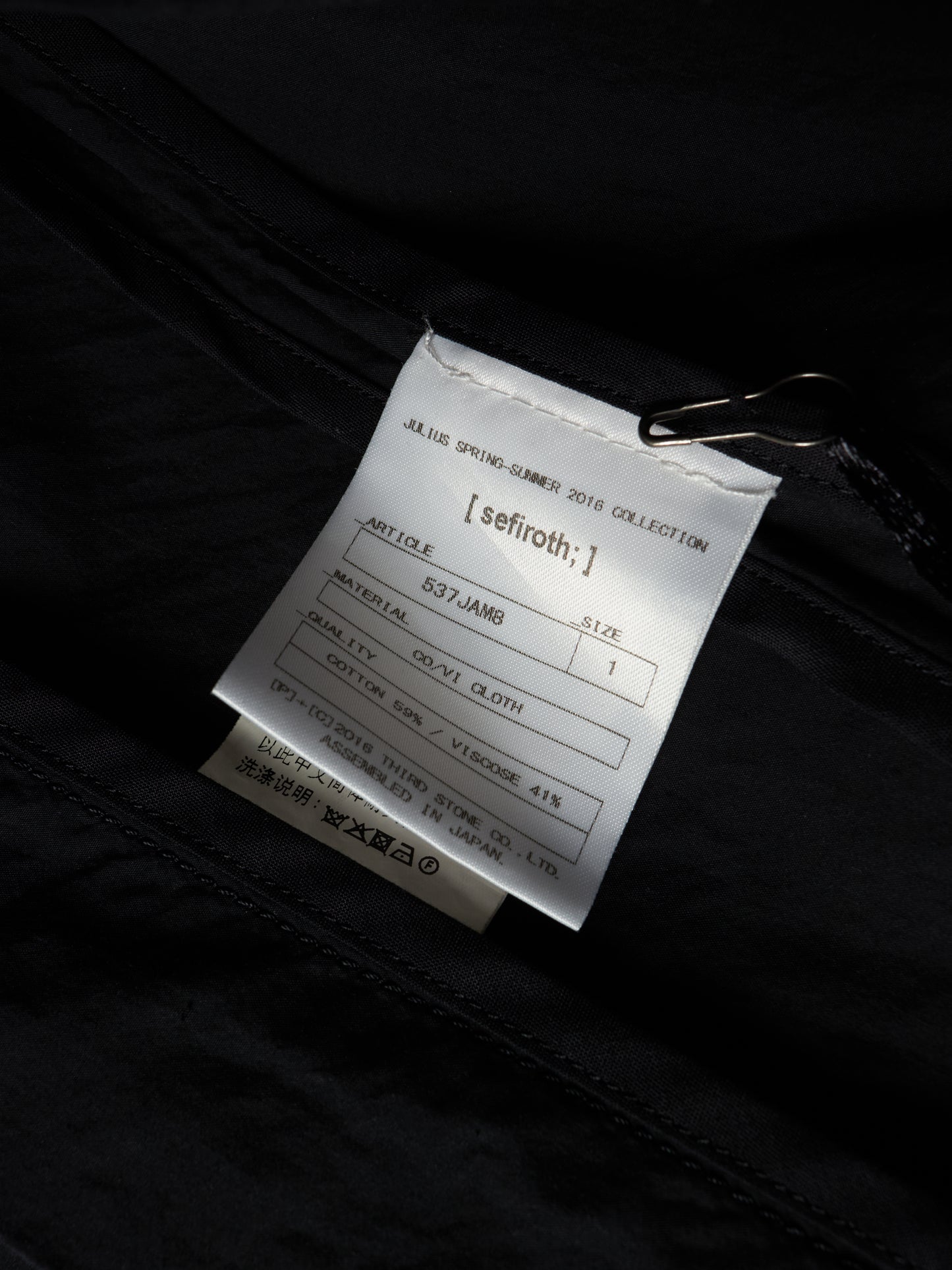 A close-up of a white product tag attached to a black garment with a metal clip, showing printed text including the brand name 'seifroth;'.