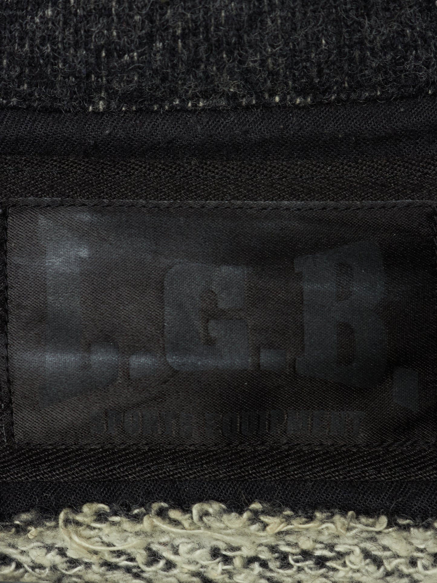 Close-up of a black woven fabric item with a dark translucent patch showing embossed letters and a textured cream and black fuzzy trim along the bottom edge.