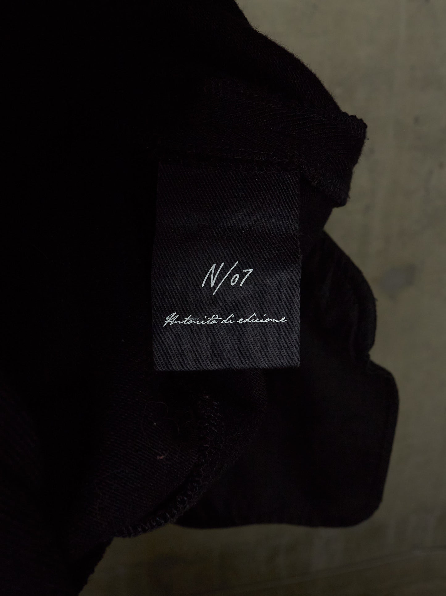 Close-up of a black garment label reading 'N/07' and 'Anticipe de chemise' on dark fabric, with a textured light gray background.