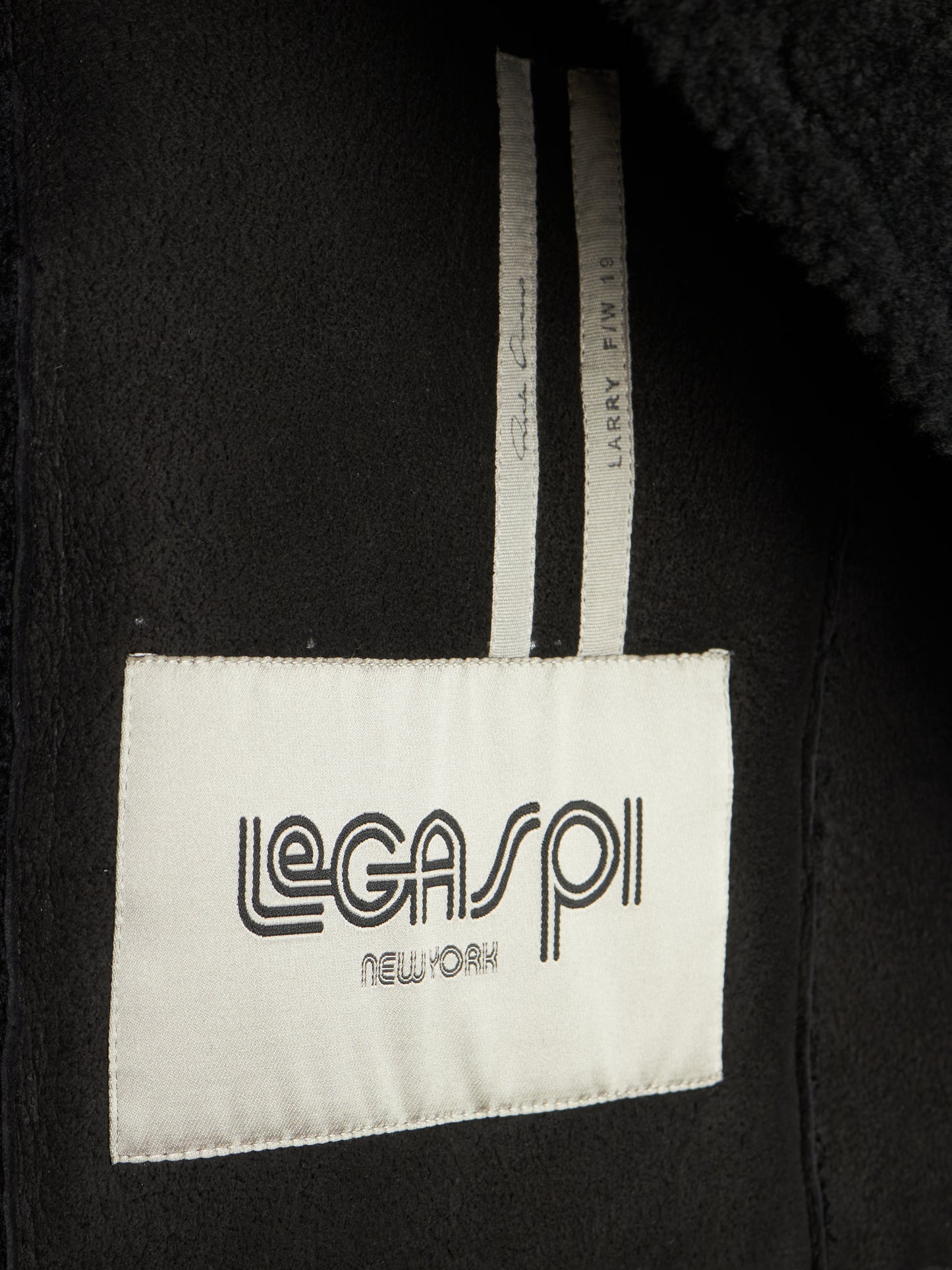 Close-up of a black garment interior showing a white sewn-in label that reads 'LEGASP1 NEW YORK' and a striped inner tag.