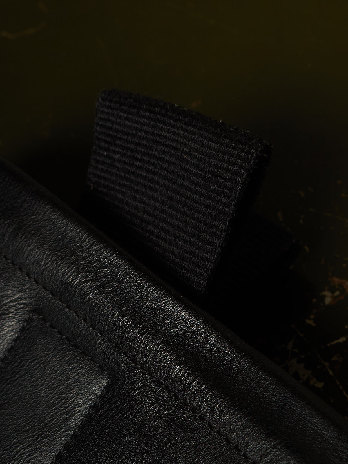 Close-up detail of a black textured leather item with a black woven strap or tab, photographed against a very dark olive-brown background.