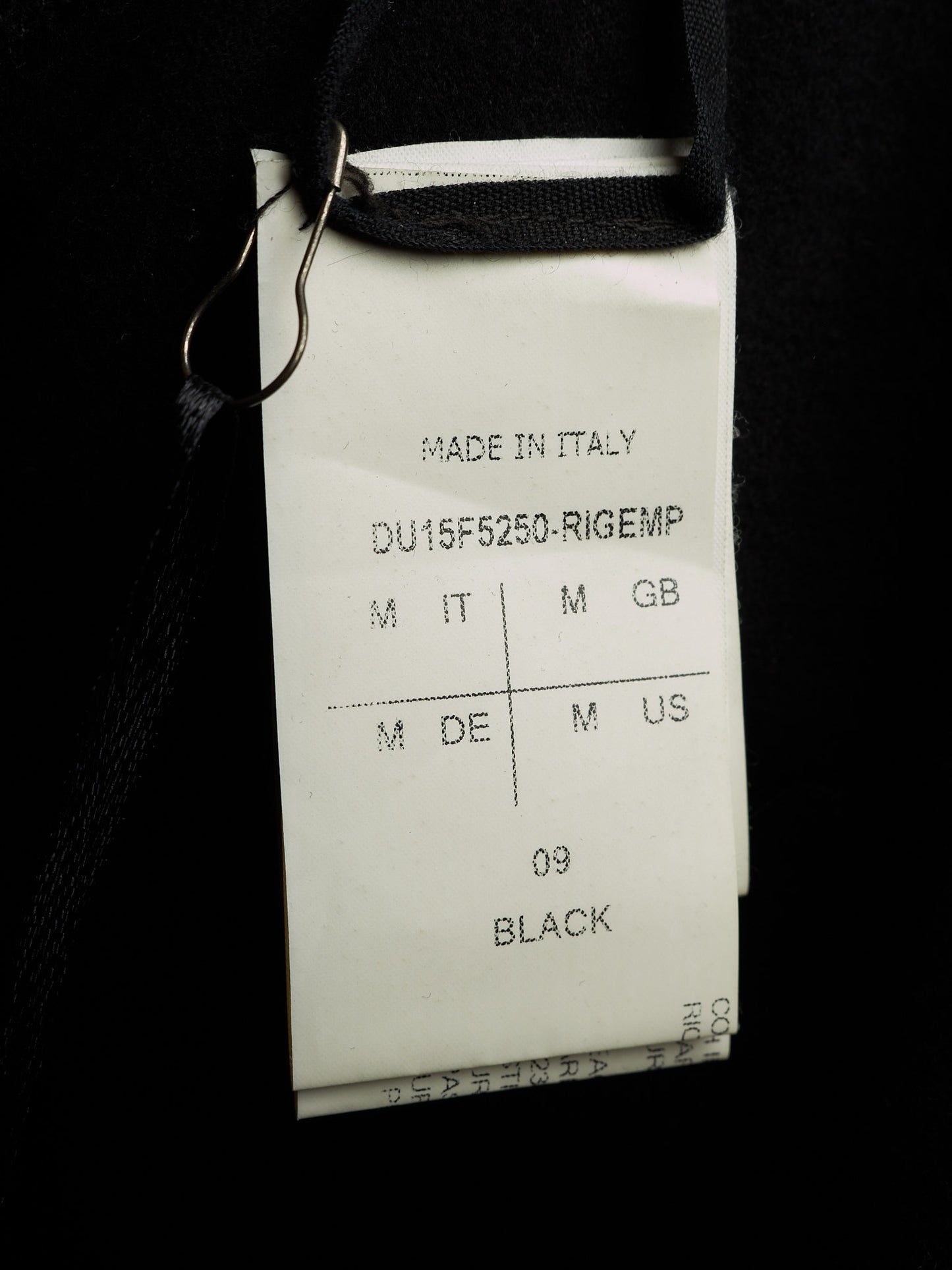 A close-up of a white garment tag attached to a black garment with a metal clip, showing text including 'MADE IN ITALY', 'DU15F5250-RIGEMP', and '09 BLACK'.