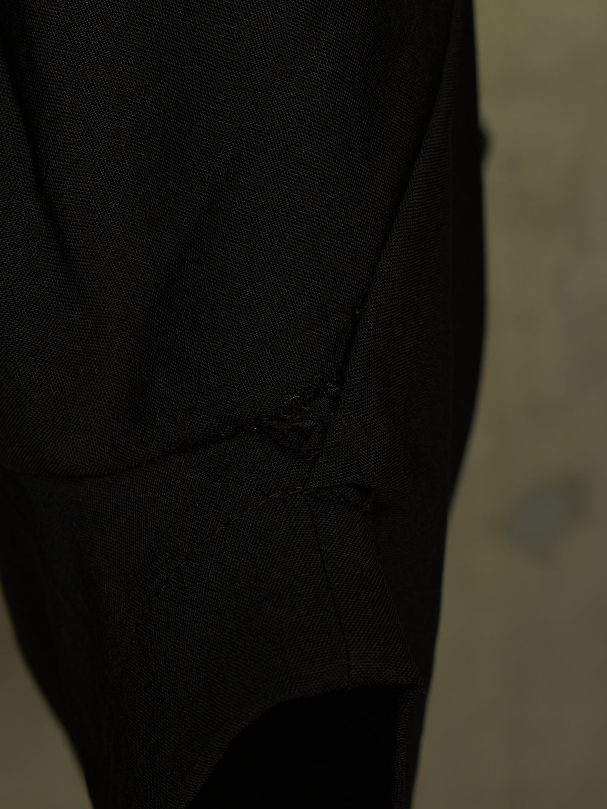 Close-up of a black textured fabric garment with a small distressed tear detail near the center, set against a dark neutral background.