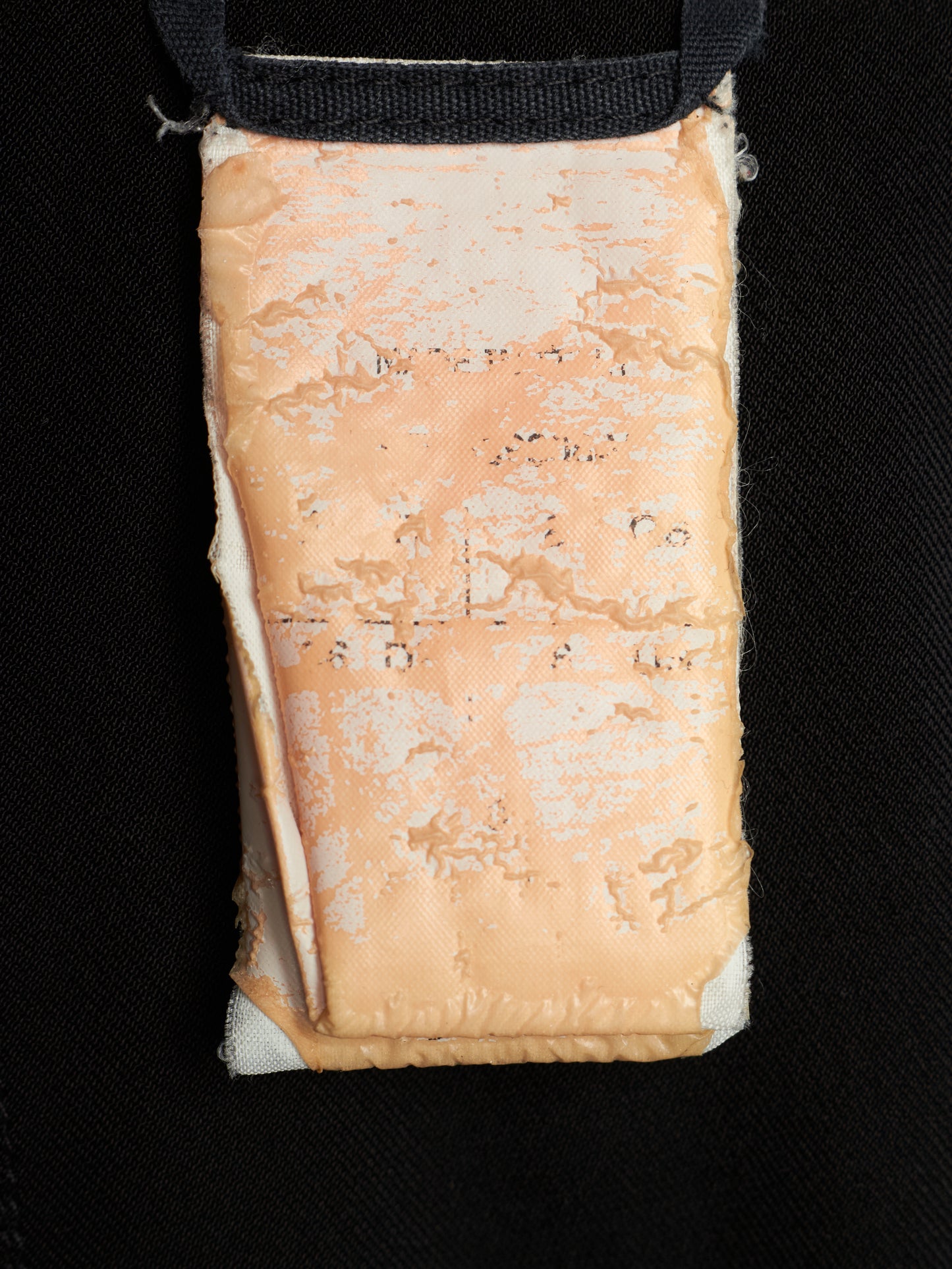 A close-up of a rectangular beige fabric pouch or pocket with a black strap at the top, shown against a black textured background.