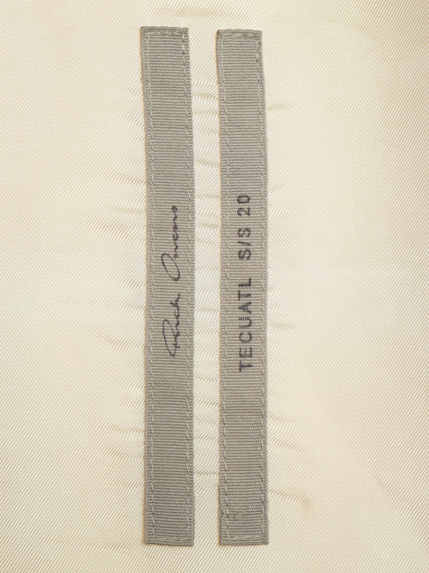 Two narrow gray fabric strips with stitched edges laid flat on a light beige textured background. One strip has a cursive signature and the other reads 'TECUATL S/S 20'.