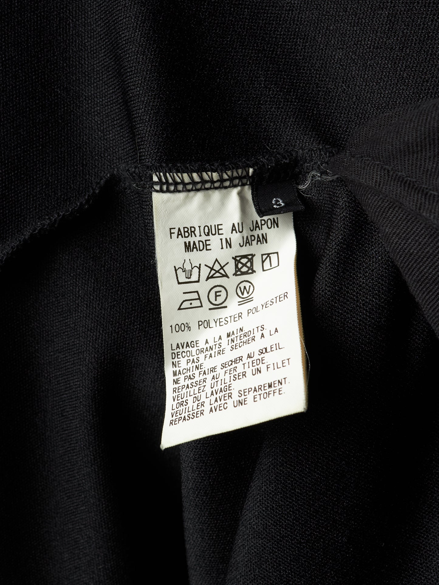 Close-up of a black garment's interior label showing 'FABRIQUE AU JAPON MADE IN JAPAN', '100% POLYESTER', and care symbols on a white paper tag.