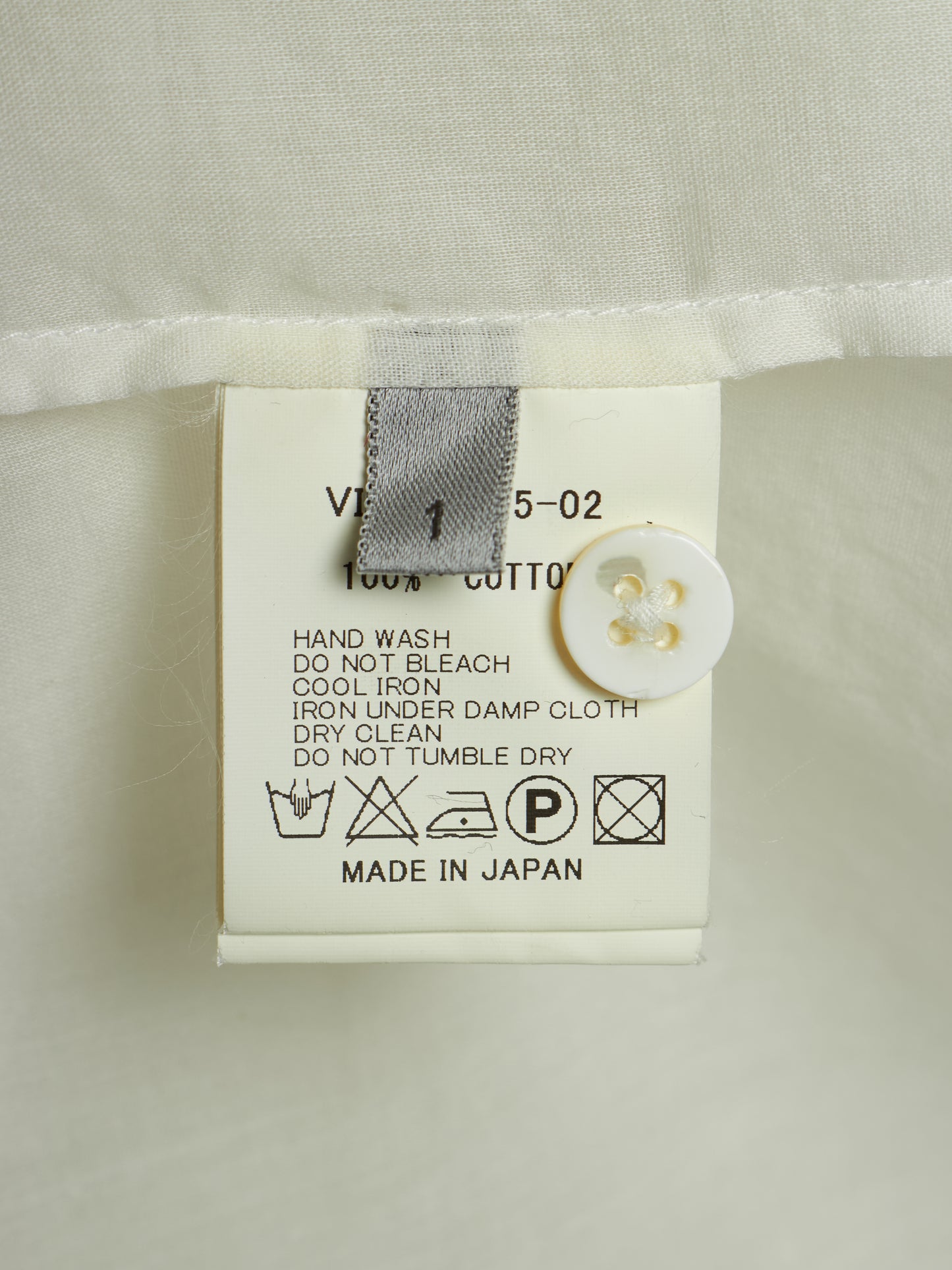 Close-up of a garment's care label attached to a white fabric hem, showing text including '100% COTTON', care symbols, and 'MADE IN JAPAN' with a white button nearby.