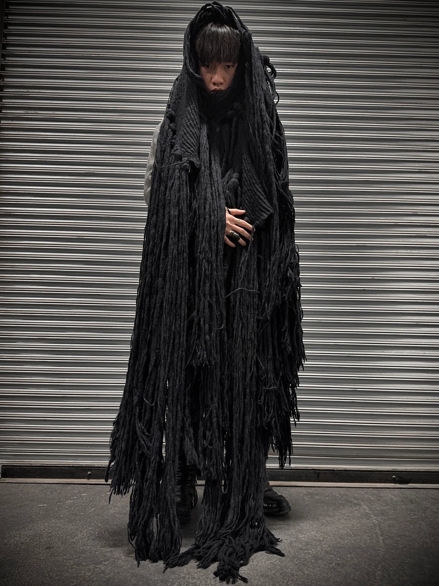 Person wearing a long, dark, textured coat in front of a metallic shutter.