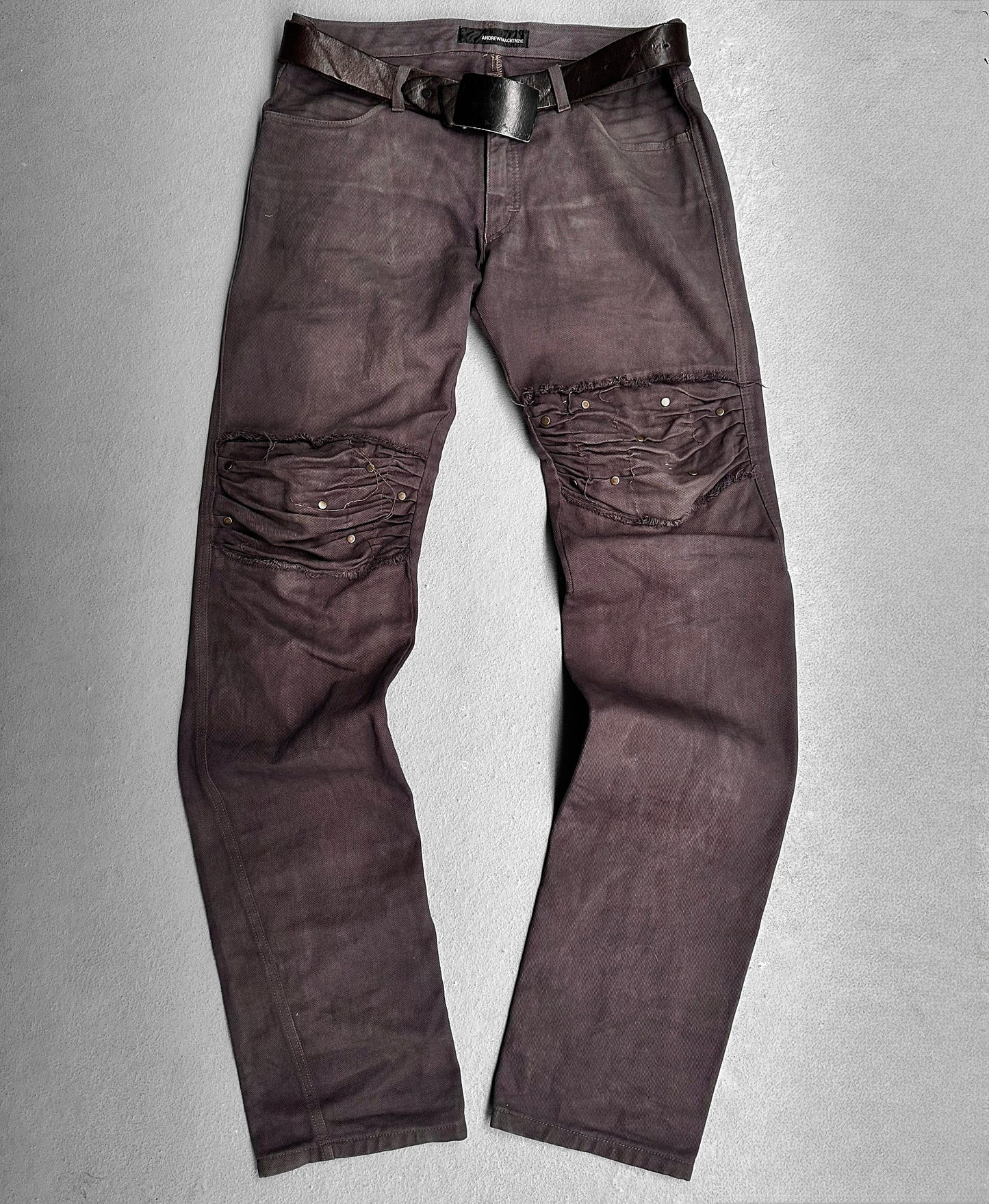 ANDREW MACKENZIE Early 00s Stud Darted Knee Denim jeans in dark wash, featuring avant-garde distressed fabric and metallic stud design on both knees, with black belt, displayed flat on a light gray surface.