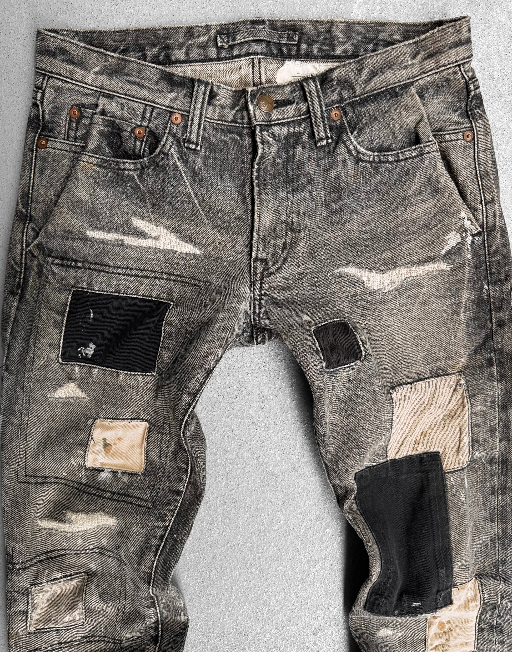 The Mihara Yasuhiro AW10 Double-pockets Repair Patchwork Selvedge Denim jeans, inspired by streetwear and the brands innovative style, feature black, beige, and striped patches and are shown laid flat on a light gray surface.