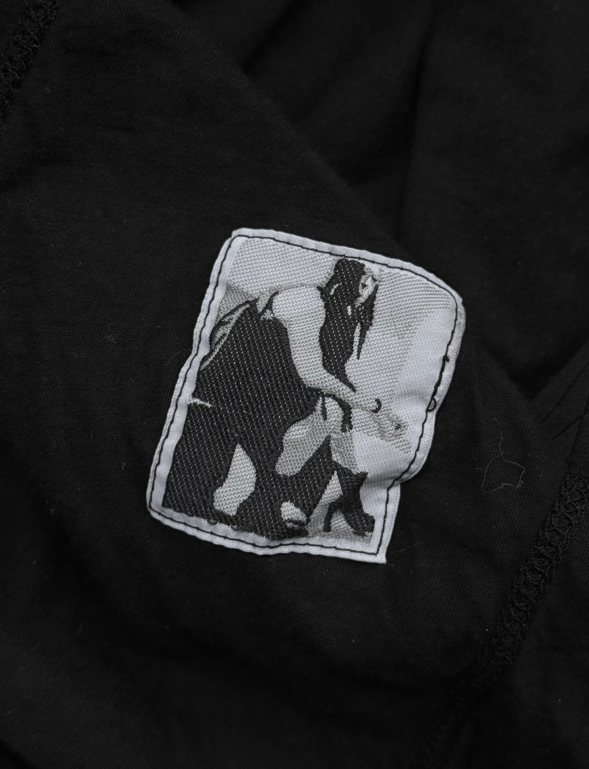 The Rick Owens DRKSHDW AW24 “PORTERVILLE” Geometric Level Long-sleeves features a black and white patch on black fabric, showing a stylized figure bending toward a small animal in pixelated, minimalist fashion.