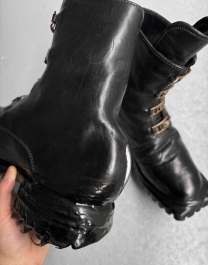 A close-up of Carol Christian Poell ‘AM/2759SP-IN CORSS-PTC’ Kangaroo Leather Buckle Drip Boots, size 8, in black with rugged soles—one boot shows creases and wear, while the other rests behind on a light gray surface.