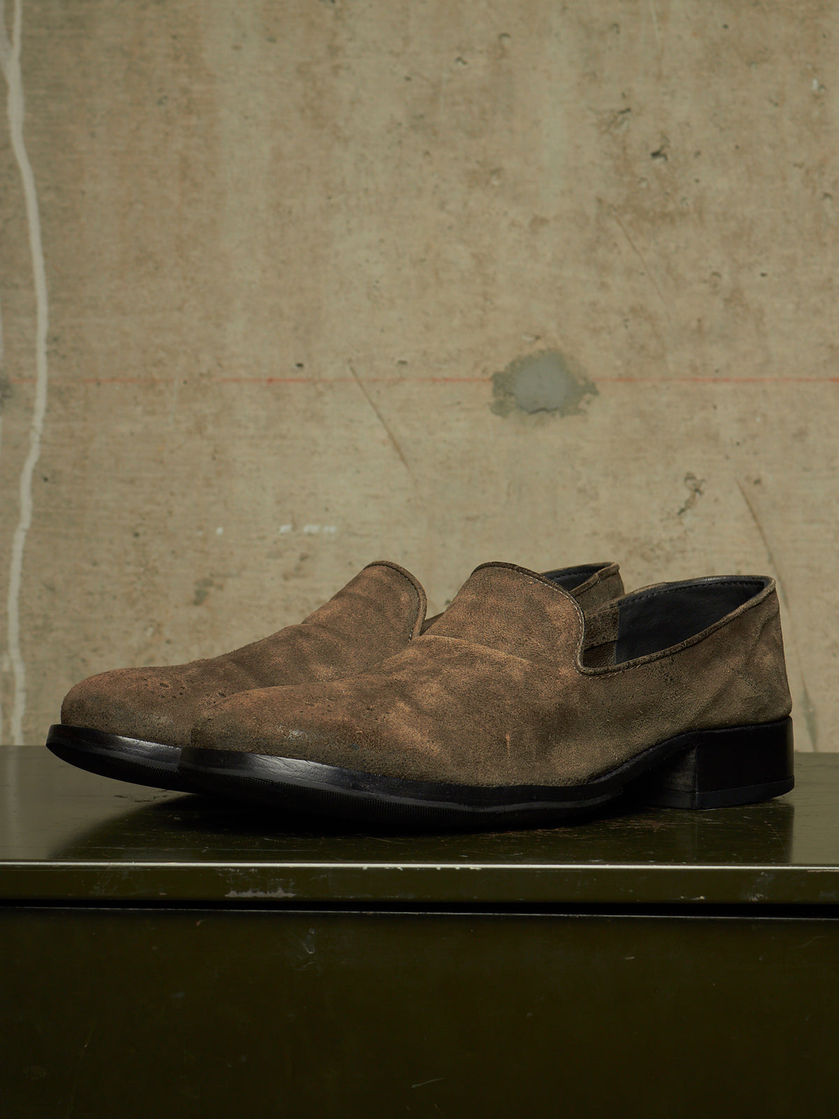 A pair of men's brown suede slip-on shoes with a low block heel, displayed on a dark green surface against a textured concrete wall.