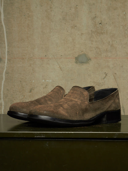 A pair of men's brown suede slip-on shoes with a low block heel, displayed on a dark green surface against a textured concrete wall.