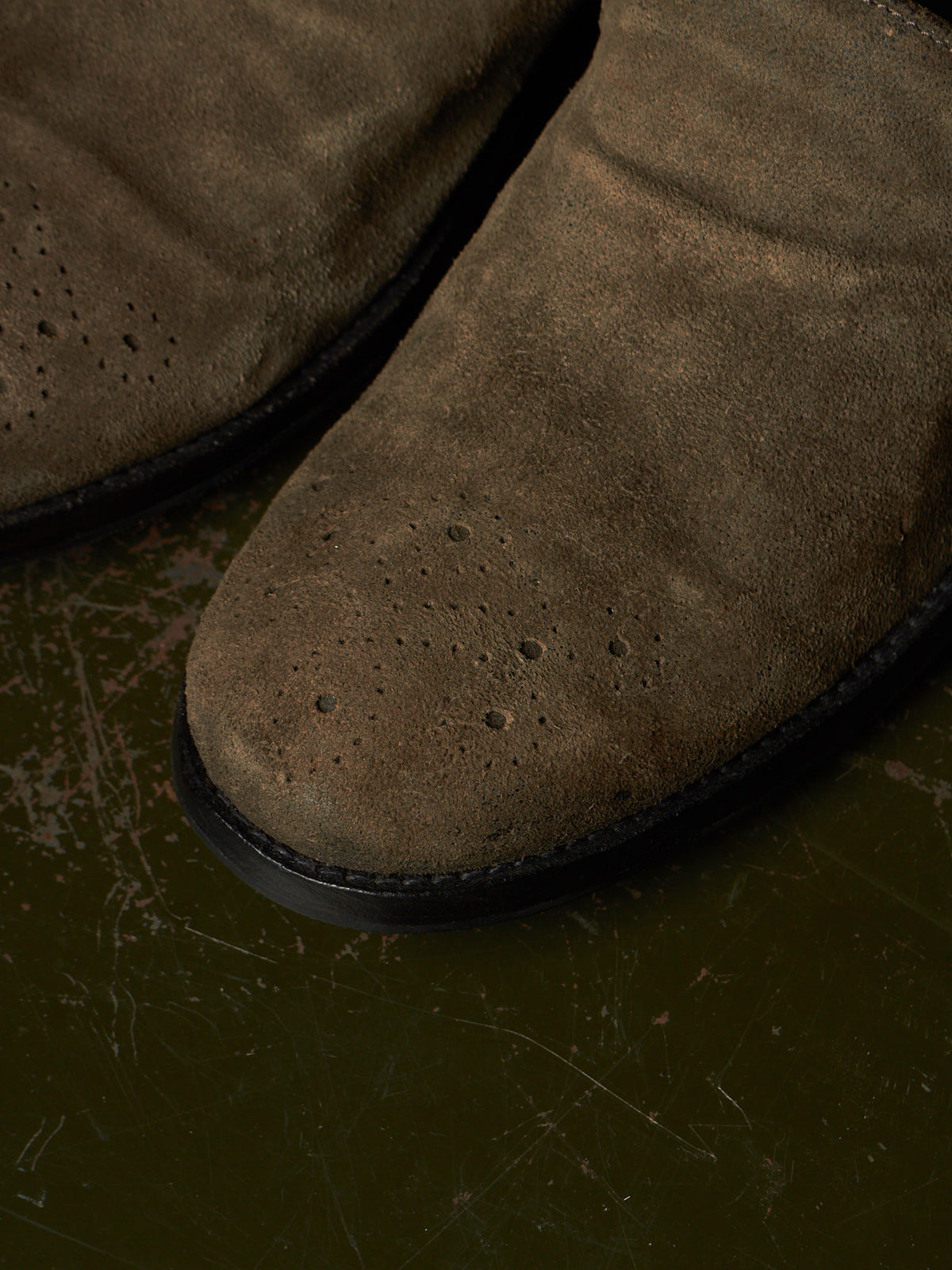 Close-up of a brown suede boot with perforated detailing on the toe, resting on a dark textured surface.