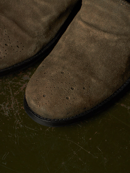 Close-up of a brown suede boot with perforated detailing on the toe, resting on a dark textured surface.