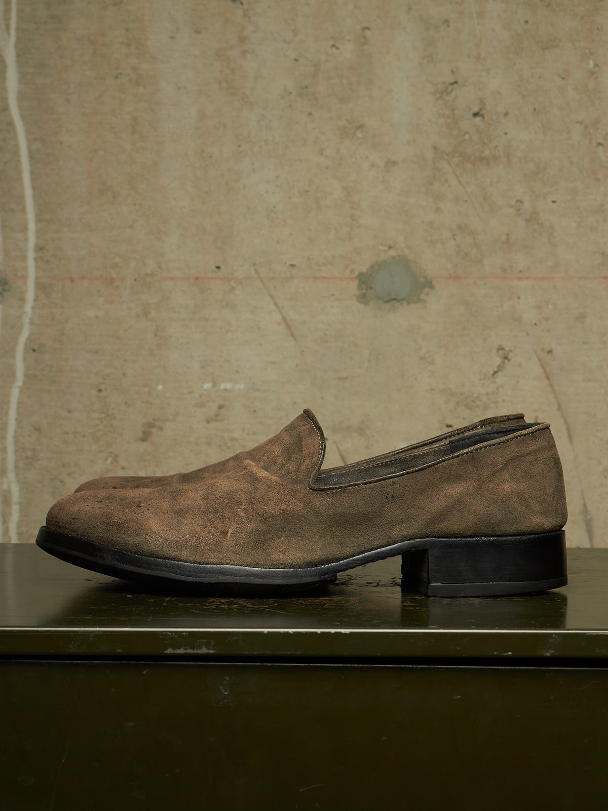 A single brown suede slip-on loafer with a low block heel, resting on a dark surface against a textured concrete wall.