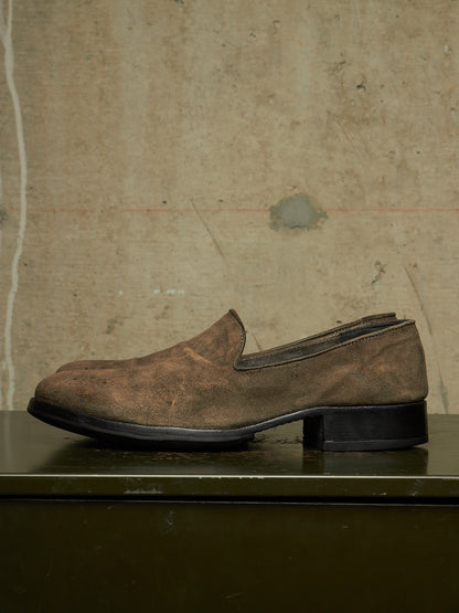A single brown suede slip-on loafer with a low block heel, resting on a dark surface against a textured concrete wall.