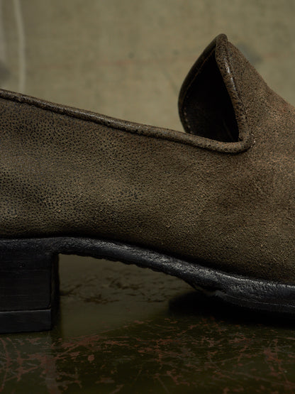 A close-up shot of a dark brown textured suede slip-on shoe, showing the grainy material, a low black block heel, and a distressed dark sole against a rustic, textured background.