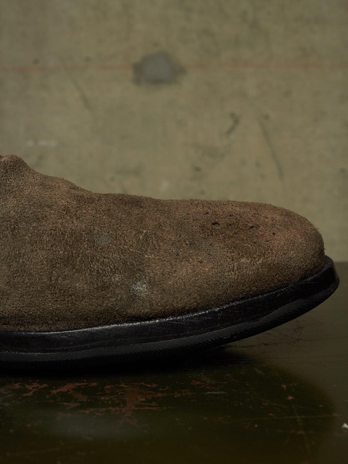 A close-up side view of a brown suede boot or shoe showing the textured upper and black sole against a dark, industrial background.