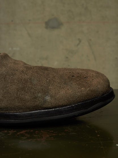 A close-up side view of a brown suede boot or shoe showing the textured upper and black sole against a dark, industrial background.