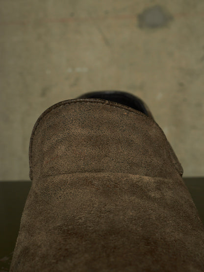 Close-up detail shot of a brown suede boot or shoe showing the textured material and a dark sole edge, set against a concrete floor background.
