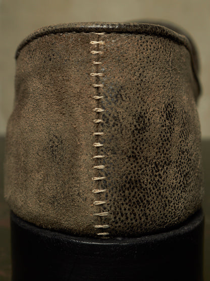 Close-up of the heel of a distressed brown leather shoe, showing a prominent vertical whipstitch and pebbled texture, with a black sole visible at the bottom.