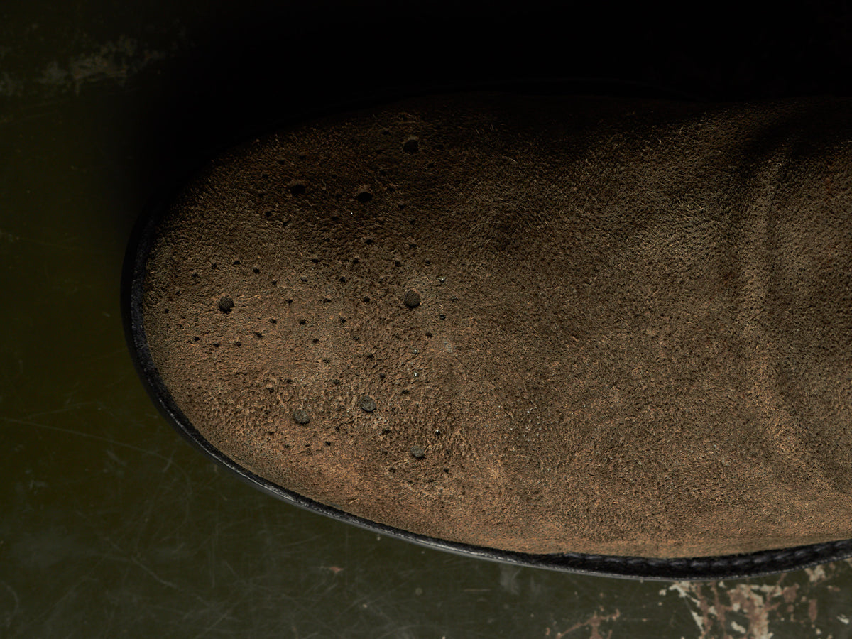 Close-up of a brown suede or nubuck boot upper with visible perforations, photographed against a dark green and black background.