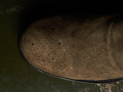 Close-up of a brown suede or nubuck boot upper with visible perforations, photographed against a dark green and black background.