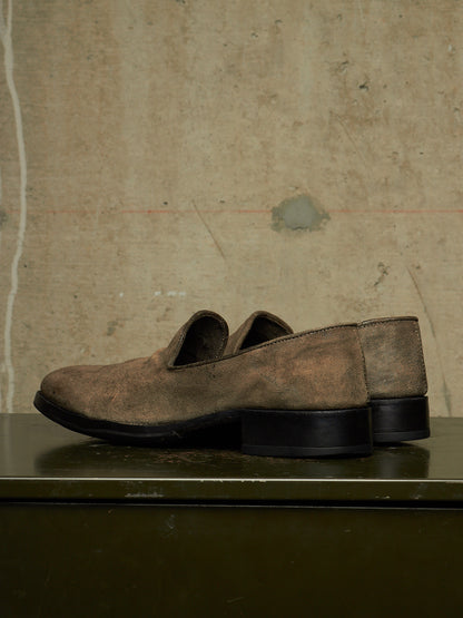 A pair of distressed brown suede slip-on loafers with black soles, displayed on a dark green metal surface against a textured concrete wall.