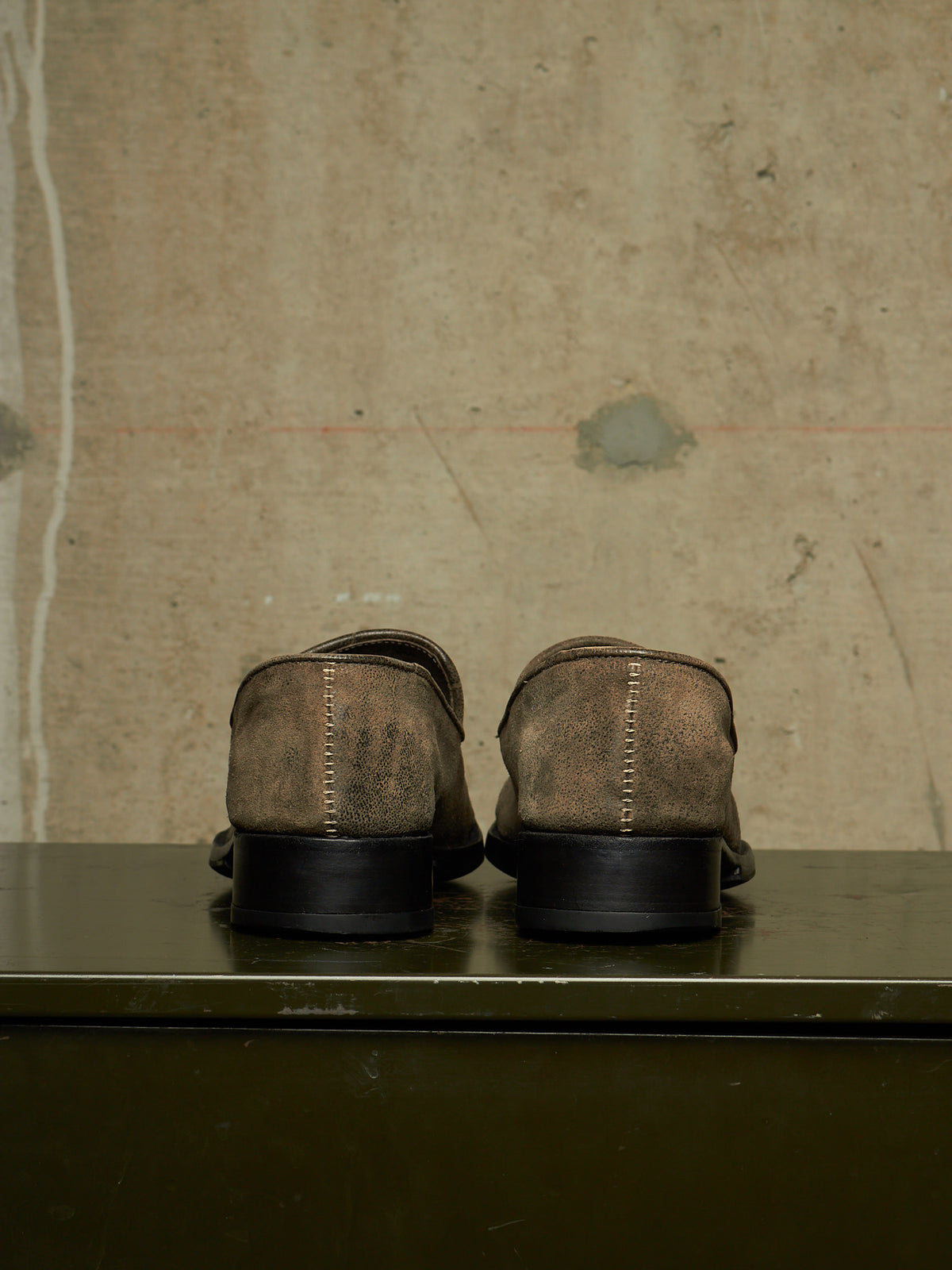 A pair of brown suede loafers shown from the back, resting on a dark green surface against a textured concrete wall.