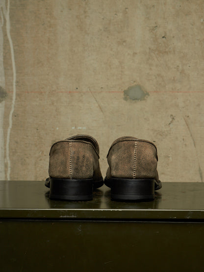 A pair of brown suede loafers shown from the back, resting on a dark green surface against a textured concrete wall.